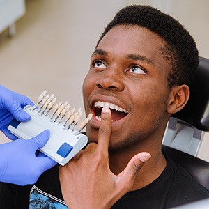 Man in dental chair pointing to teeth with shade guide held close to his smile
